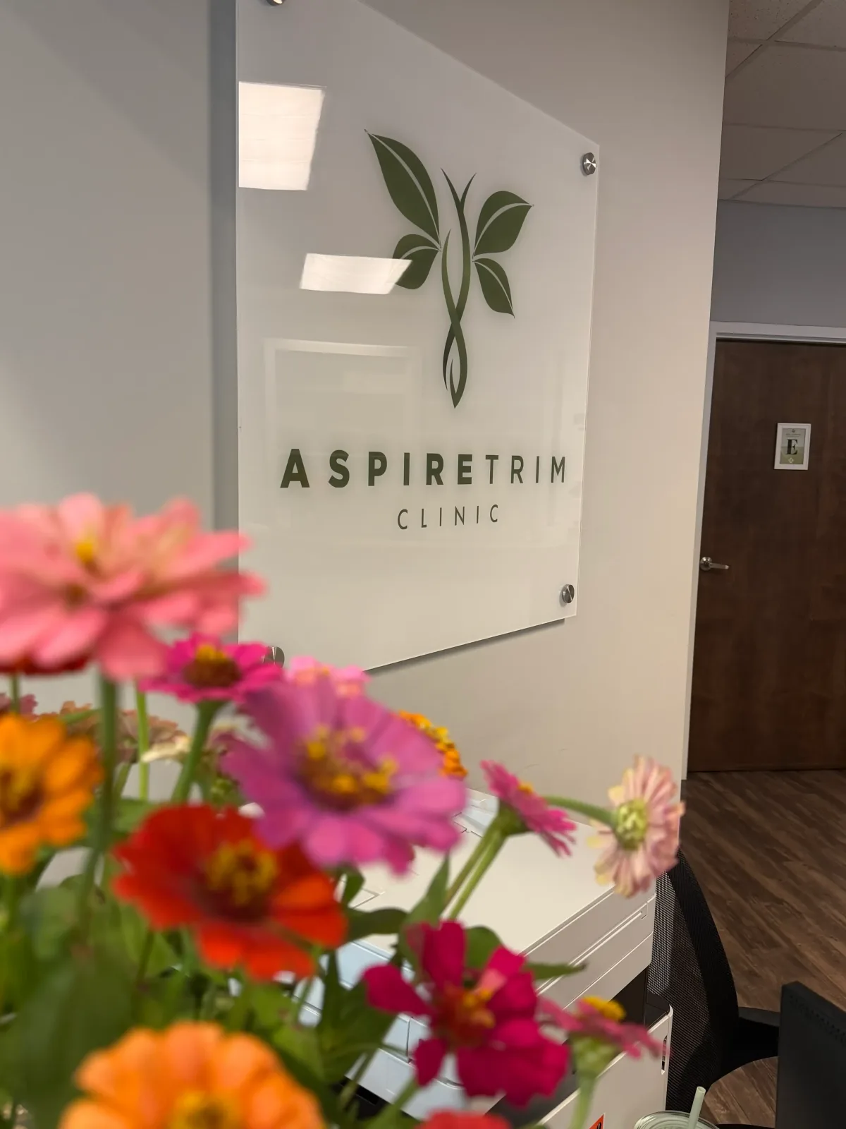 Aspire Trim Clinic logo sign behind fresh zinnias at the front desk of the Matthews, NC clinic